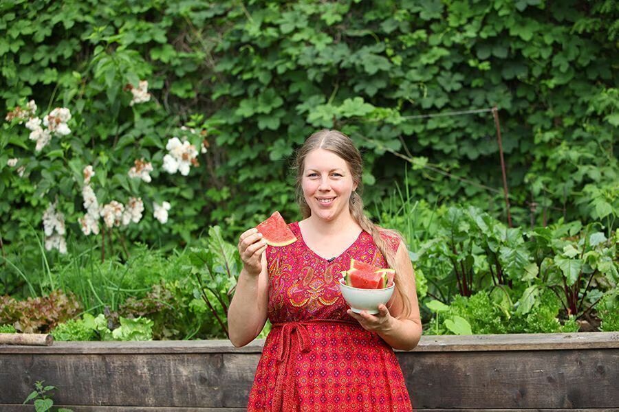 Jen steht lachend mit einem Stück Wassermelone in der Hand vor den Hochbeeten im Hofgarten