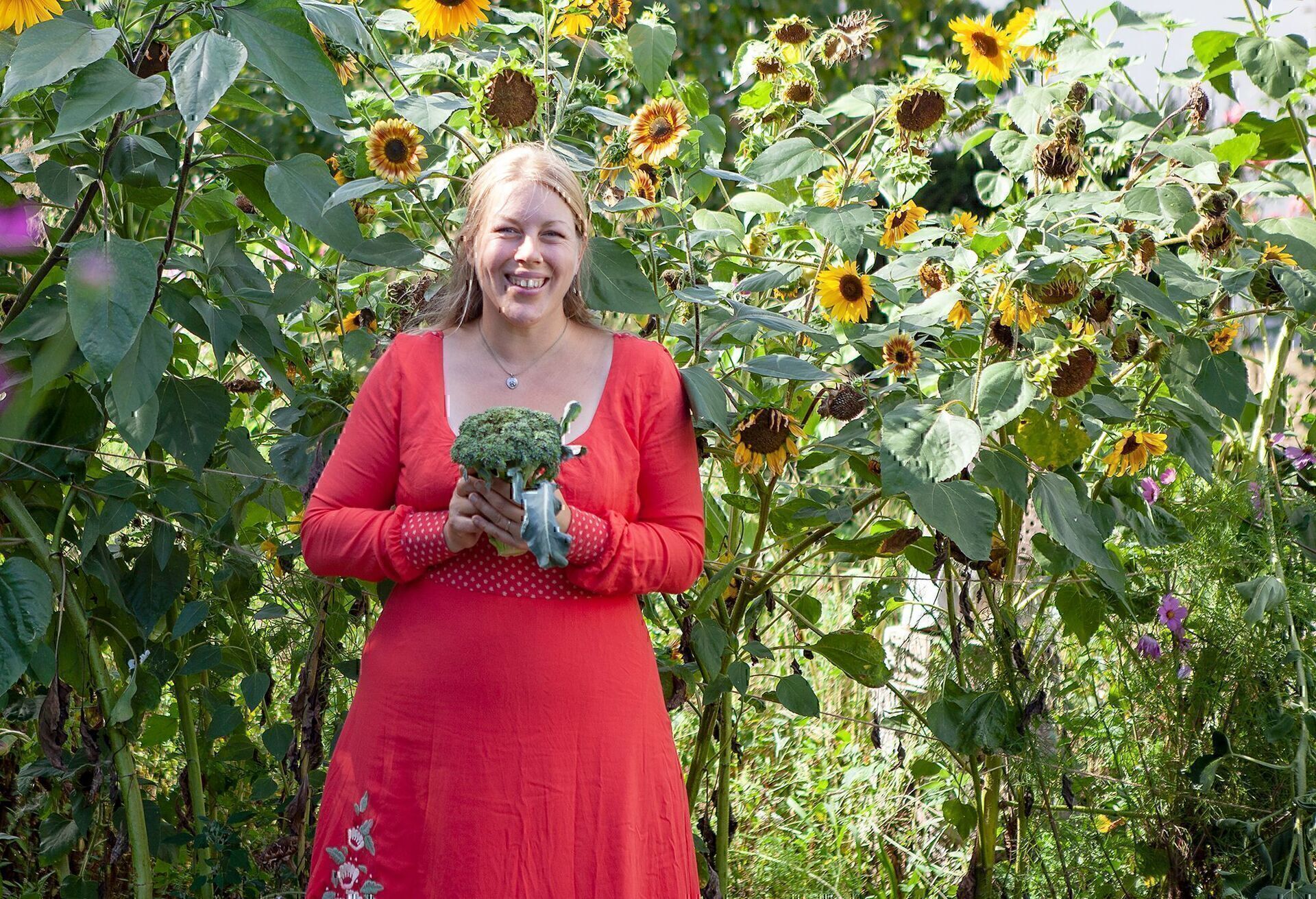 Jen im roten Kleid hält einen frisch geernteten Brokkolikopf vor blühenden Sonnenblumen