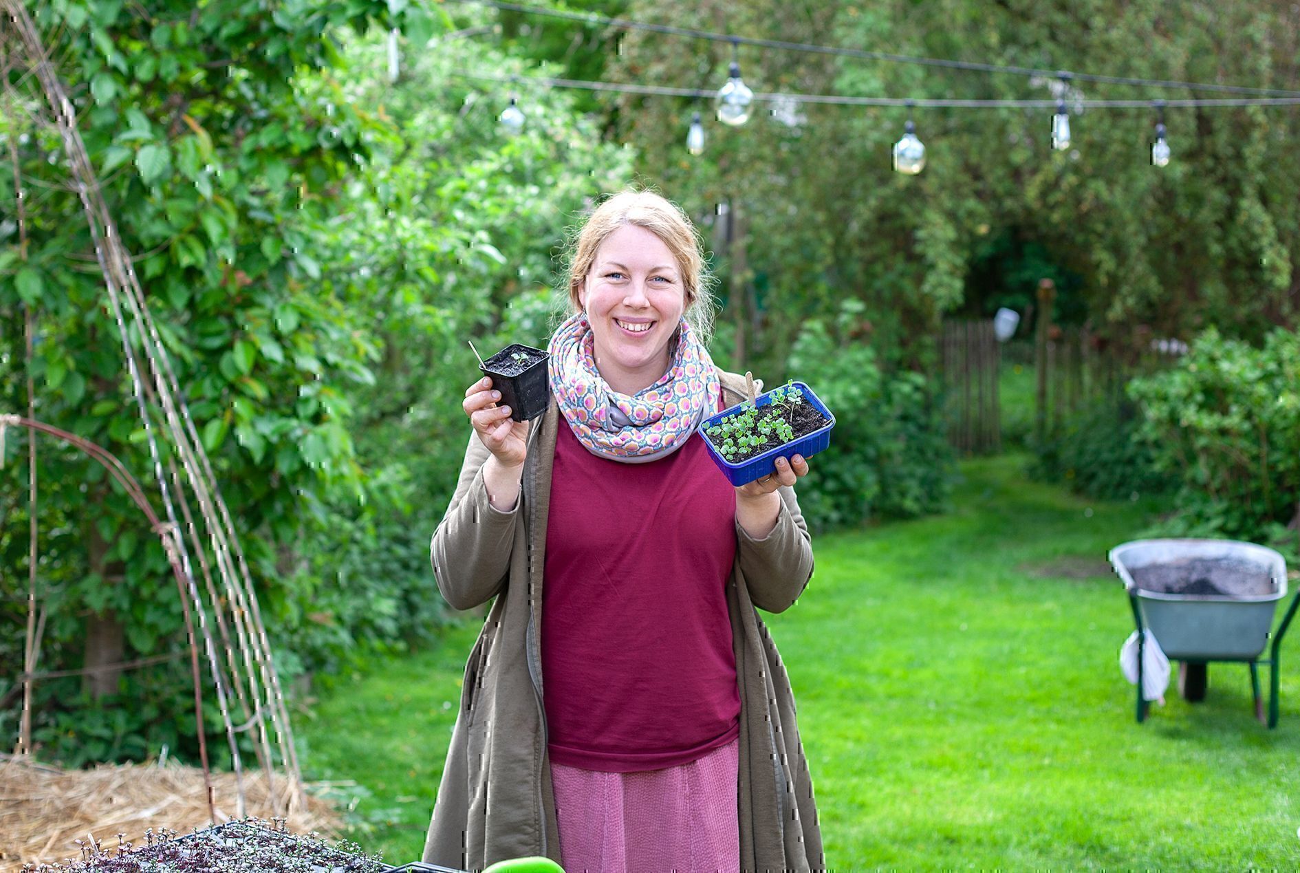Jen steht im Garten und hält ein Töpfchen und eine Schale mit jungen Wirsingpflanzen