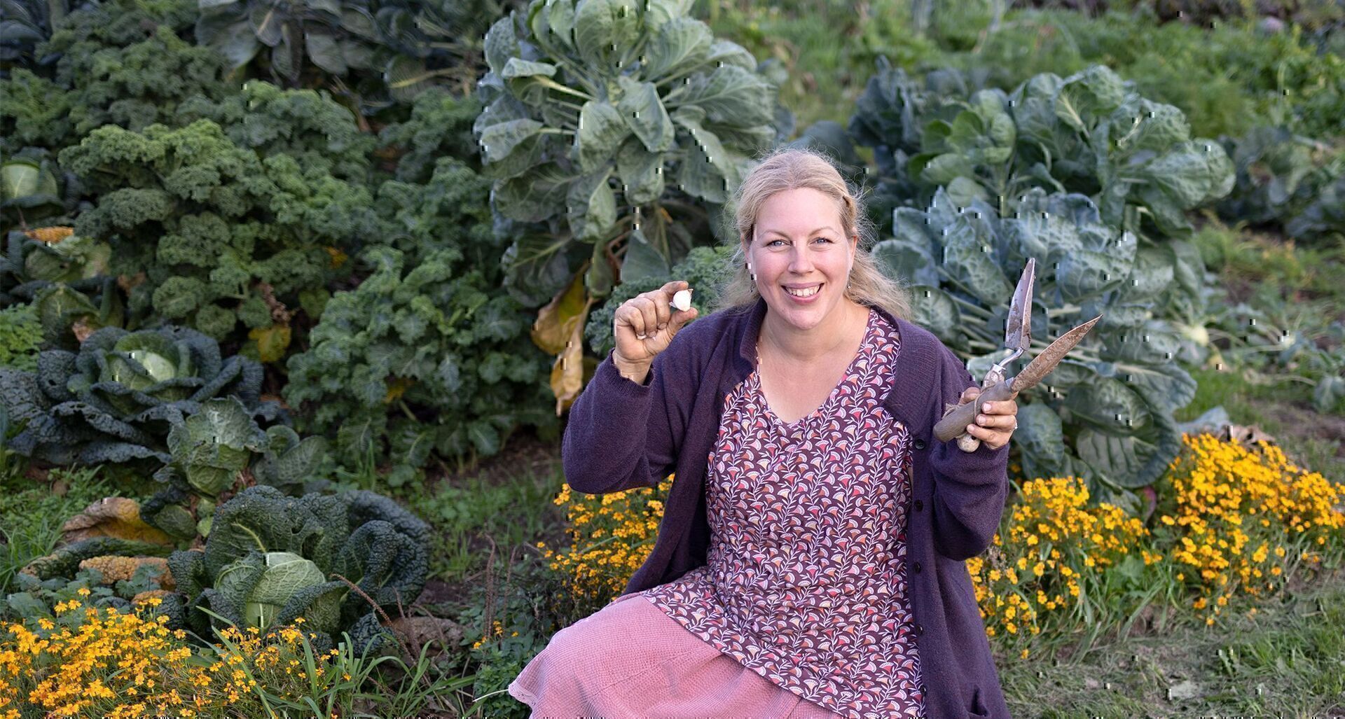Jen hockt vor Kohlpflanzen und Tagetes und hält eine Knoblauchzehe und ein Pflanzholz in die Kamera