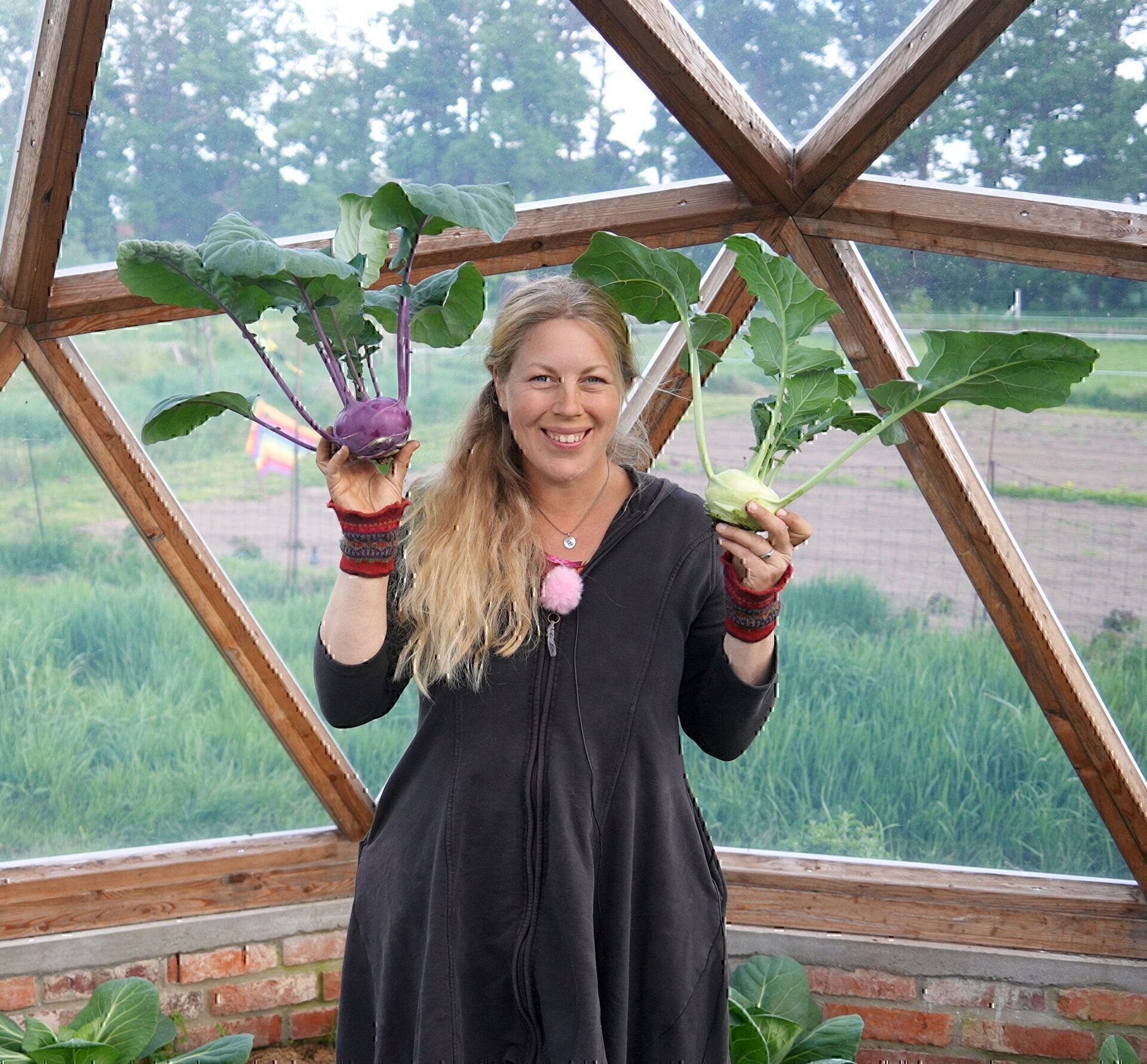 Jen hält im Kuppelgewächshaus einen lila und einen weißen Kohlrabi mit Blättern in die Höhe
